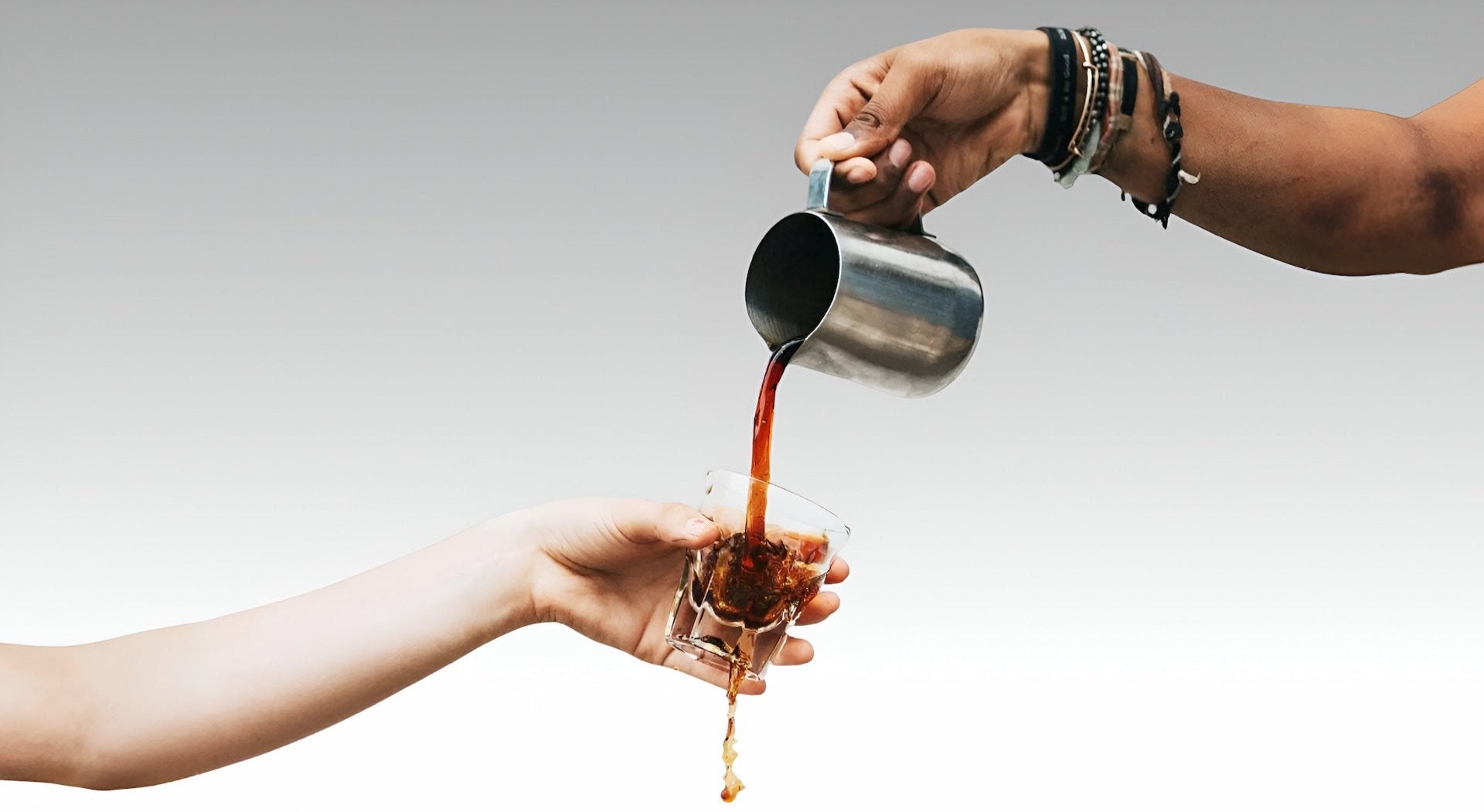 Two hands pouring coffee from a metal pitcher into a glass, with the coffee mid-stream against a white background.